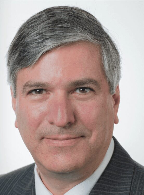 Professional headshot of a middle-aged man with gray hair in a dark pinstripe suit against a light background.