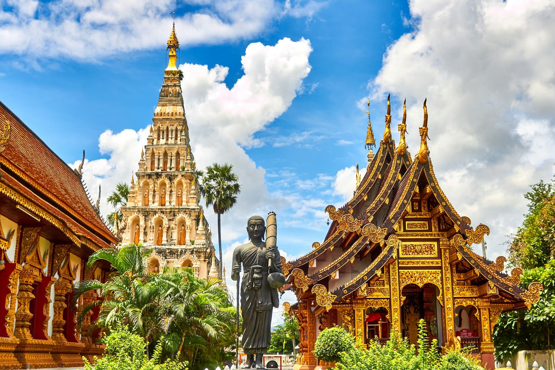 Ornate Thai Buddhist temple complex with gilded roofs, a tall white pagoda, and a standing Buddha statue under a blue sky.