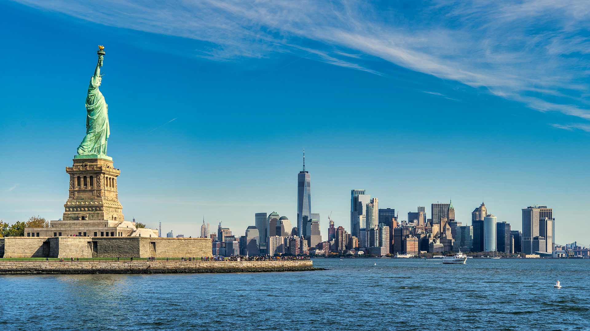 Statue of Liberty on a stone pedestal with NYC skyline across a harbor under a blue sky.